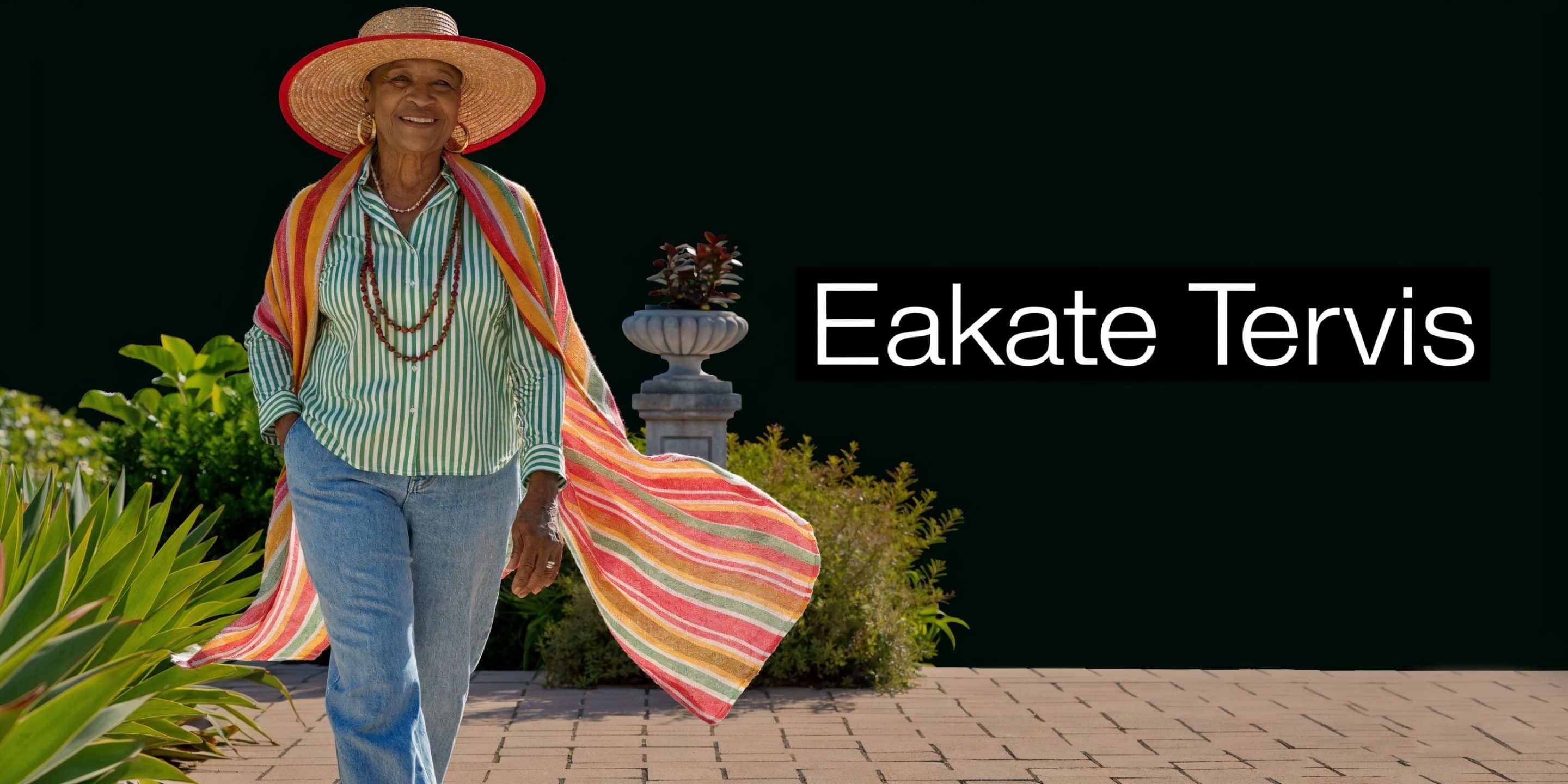 Naeratav eakas naine jalutab väljaspool, kandes suvemütsi ja värvilist triibulist salli, luues rõõmsameelse ja stiilse välimuse.